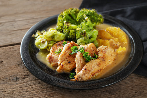 Chicken fillet in orange sauce with broccoli leek vegetables on a black plate on dark rustic wood, low carb diet dish, copy space, selected focus, narrow depth of field