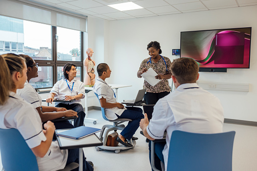 Group of medical students in a classroom in a hospital studying for their medical degree. They are at a healthcare facility in Newcastle Upon Tyne, North East England. The students are all wearing their student nurse uniforms. A female teacher is supervising the students, handing out documents.