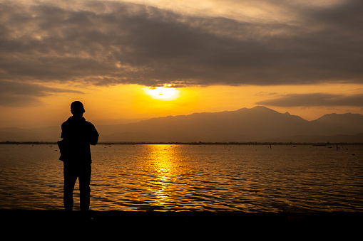 A young man in black is taking pictures of the sunset behind the mountain. And there are bright colored lights reflected on the water surface as a beautiful background.