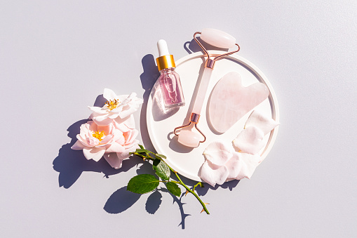 Bottle of Cosmetic Rose Oil, Quartz Massager, Gua Sha Scraper on White Ceramic Tray for Home Massage and Self-Care. Top view