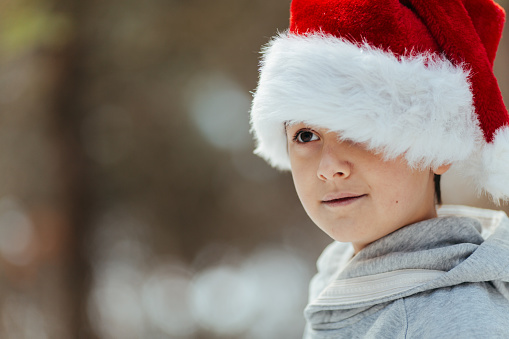 Little boy with Santa hat staning on a snow in a winter forest