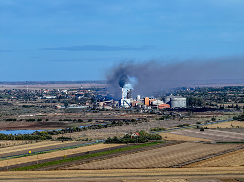 Patchwork farmlands, reminiscent of simpler times, cradle a small village where life seemingly moves at a gentler pace. Yet, the narrative shifts dramatically towards the presence of a behemoth industrial setup. Its structures, accompanied by the unmistakable sight of rising smoke, signal the relentless march of industry, standing in sharp relief against the backdrop of nature and tradition.