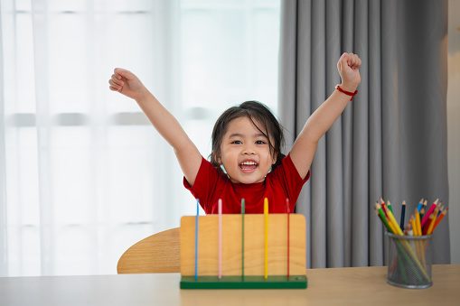 Young cute Asian baby girl wearing red t-shirt is learning the abacus with colored beads to learn how to count on the table in the living room at home. Child baby girl development studying concept.