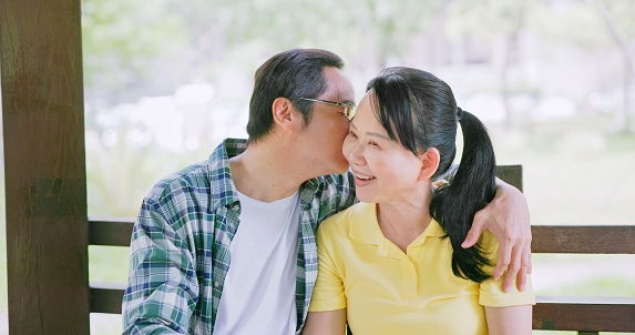 Closeup view of asian elderly couple happily sit on public gazebo bench chat and talk in the park - he is kissing his wife cheek