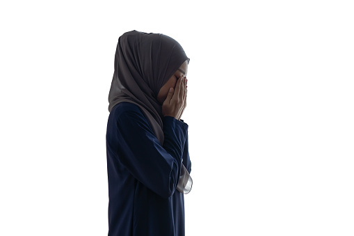 Beautiful Asian Muslim woman praying to god, hands up, wearing traditional headscarf. Pretty female Islamic near window.