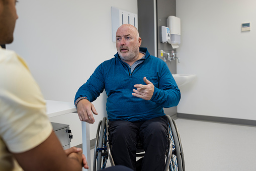 An over the shoulder view of a male doctor sitting in his clinic with a mature man who is a wheelchair user. He is explaining his concerns and discussing things with the doctor. They are sitting in clinic in Newcastle upon Tyne in the North East of England.