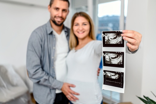 Husband is embracing his pregnant wife next to the window in their new home.