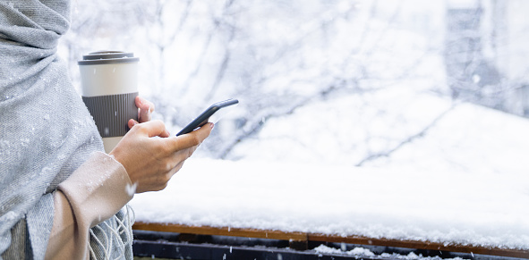 Close up hands of a woman  with smartphone and cup of coffee.  Winter landscape with snowfall in the background
