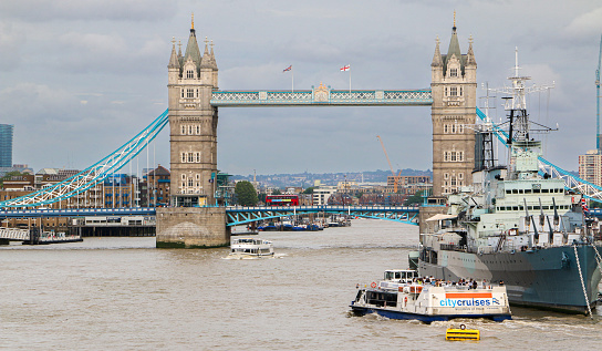 View of the Iconic Tower Bridge as a tour boat cruises past the HMS Belfast ship moored on the banks of river Thames - London, UK