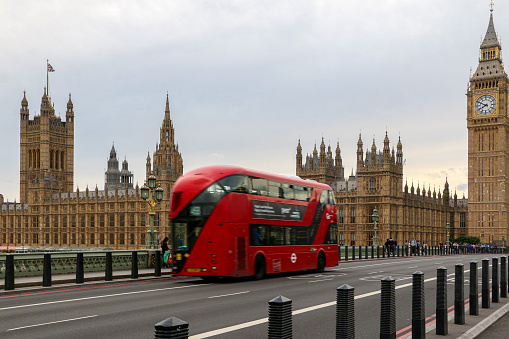 British London iconography, red double decker bus crosses Westminster Bridge with sight of Big Ben and the Houses of Parliament in background - London, UK