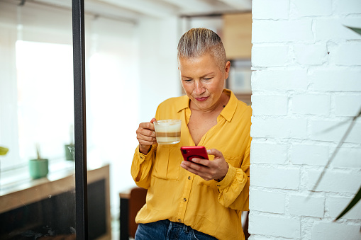 Happy woman at home drinking coffee while using her phone