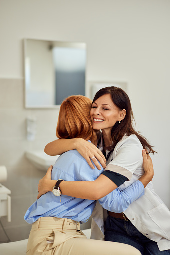 A brunette female doctor hugging a female patient, sitting on the bed, smiling.