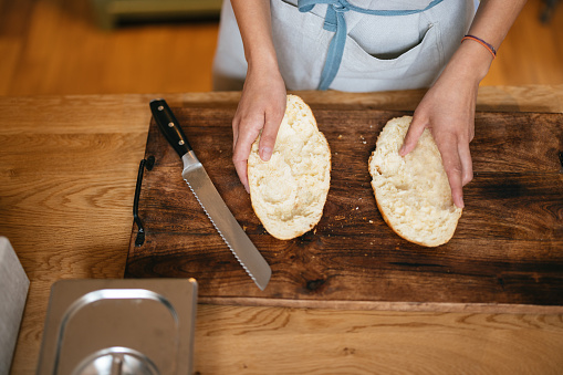 Unrecognizable woman standing by the kitchen counter holding two halves of the bread. There is a bread knife which she used to cut it in half.