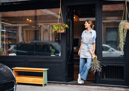 Happy woman in apron holding a bouquet of lavender and looking away while standing at the store doorway.