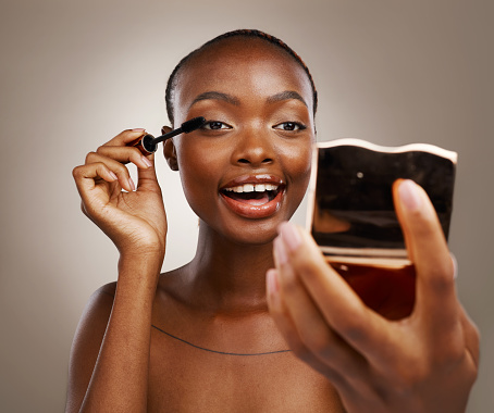 Black woman, mascara with brush and mirror, makeup and shine with beauty isolated on brown background. Eyelash extension, wand and skin glow with cosmetology, transformation and wellness in studio