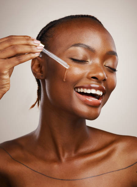 Happy woman, skincare and serum on face in studio for beauty, dermatology or collagen on brown background. African model, facial cosmetics and liquid dropper of hyaluronic acid, essential oil or glow stock photo