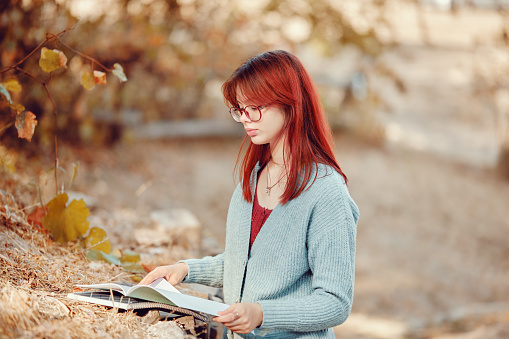 Red-haired student girl is studying outside, putting notebooks on the grass.