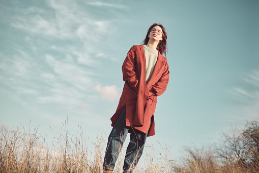 Teenage girl in a red coat stands against the sky with her hands in pockets.