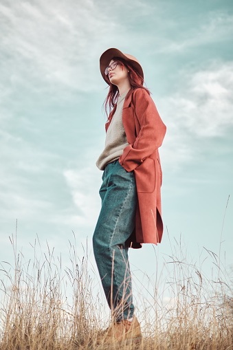 Confident teenage girl in red coat and hat standing against the sky with hands in pockets and looking away. Low angle view.