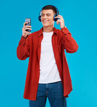 Man, smile and headphones for audio on phone in studio by blue background. Asian person, searching and streaming a song, radio and music or funny podcast on smartphone, technology and sound for joy