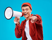 Portrait, bullhorn and young man in studio pointing for an announcement or speech at a rally. Happy, smile and male activist on stool with megaphone for loud communication isolated by blue background