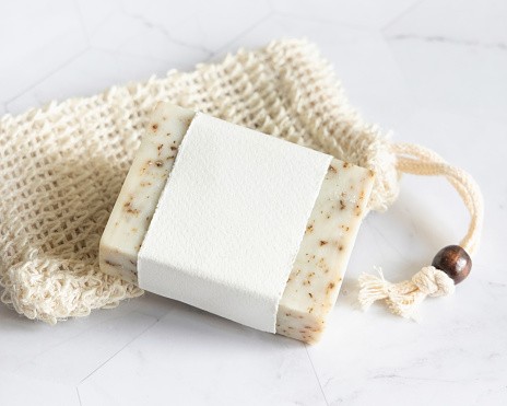 Beige handmade soap bar with blank label on sisal soap saver bag on marble table close up, mockup, copy space. Natural herbal products for face and body care.