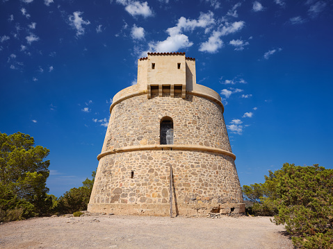Wide-angle view of Torre d'en Valls, also known as Torre de Campanitx, an 18th-century watchtower located on the eastern coast of Ibiza. The dazzling bright light of a Mediterranean summer afternoon, a deep blue sky, picturesque clouds, lush bushes. High level of detail, natural rendition, realistic feel. Developed from RAW.
