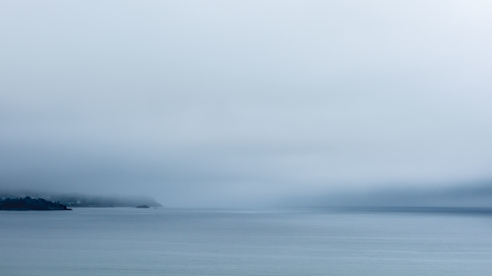 Mysterious atmosphere in Brittany with thick mist, gray skies, and the Breton coast visible on the horizon. The sea in the foreground is remarkably calm, reflecting gray and blue tones.