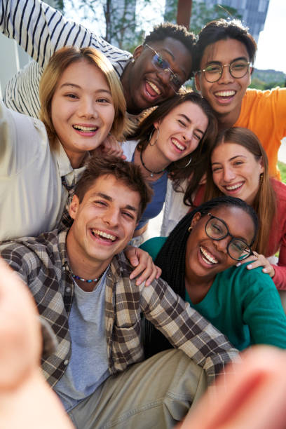 Multiethnic classmate taking a selfie outside of the university, looking at camera cheerfully. Happy group of multiethnic classmate taking a selfie together outdoors in the university campus, looking at camera cheerfully raising arms, celebrating last day. Vertical photo. teens interacting photos stock pictures, royalty-free photos & images