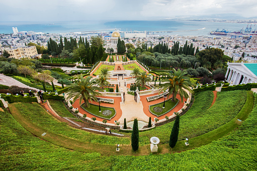 Bahai Gardens (Bahai Bab) in Haifa city on  steep slope of Carmel  Mount.  Wonderful views of the city and the Mediterranean Sea and port with machinery cranes\nBlurred motion people: gardener and tourists