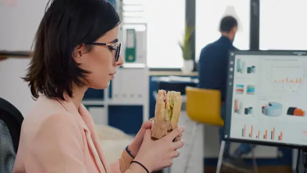 Entrepreneur woman eating tasty sandwich during takeout lunchbreak working at financial graphs in startup business company office. Fast food order paper bag delivered at workplace