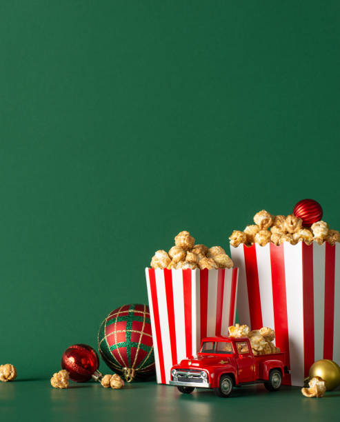 istock Opt for stay-at-home holiday experience with delivery idea. This side vertical view image captures tabletop featuring popcorn, festive baubles, tiny car on green wall backdrop, ideal for movie advert or promo 1765346675