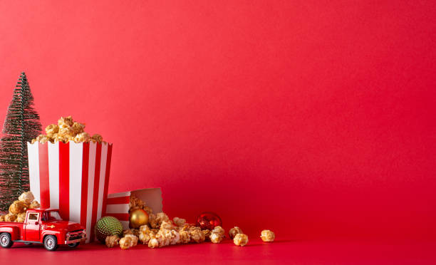 istock Create perfect Christmas movie night at home with delivered popcorn. Side-view image featuring popcorn boxes, balls, miniature vintage car, fir tree set against red wall, ideal for promotional content 1765346858