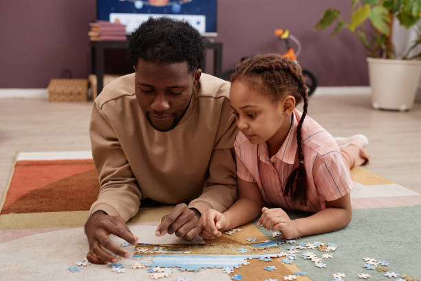 istock Black little girl solving jigsaw puzzle laying on floor with father 1765380122