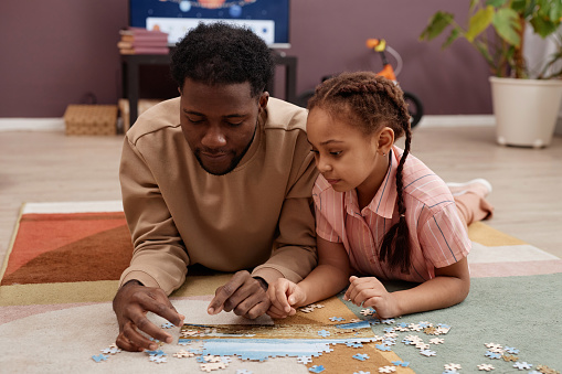 Portrait of Black little girl solving jigsaw puzzle while laying on floor with father and playing together at home