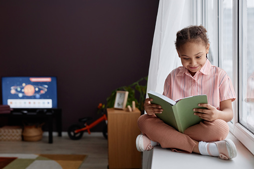 Full length portrait of cute Black little girl sitting on window and reading book in pink room interior, copy space