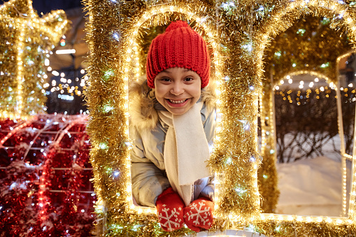 Front view portrait of smiling Black girl looking at camera through window playing with outdoor Christmas decorations