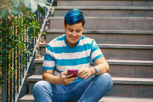 In a serene urban setting, a non binary individual with striking blue hair and a stylish striped shirt is engrossed in their phone. The scene evokes the blend of nature and technology in modern life.
