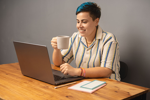 A modern and stylish genderfluid woman with blue hair sits at her home office, sipping coffee and working on her laptop, showcasing a contemporary work-from-home setting.
