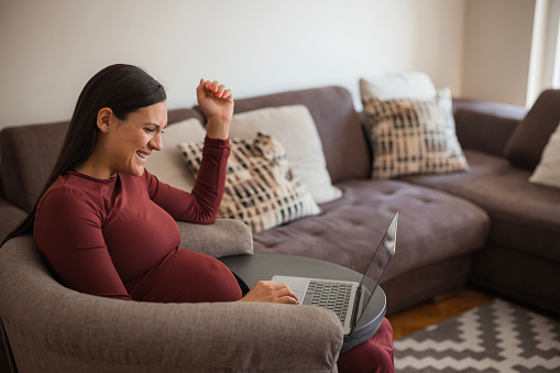 Happy pregnant woman laughing and using laptop when sitting in a sofa at home