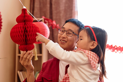 Portrait of cute Asian little girl hanging red paper lanterns at home with father preparing for Chinese New Year celebration, copy space
