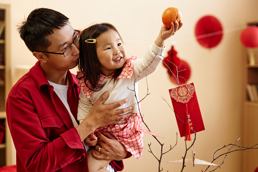 Waist up portrait of happy Asian girl holding tangerine while celebrating Chinese New year with father at home, copy space Translation Have overflowing abundance hundred years