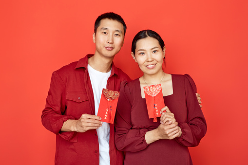 Waist up portrait of young Chinese couple holding red hongbao envelopes with good luck inscription and smiling at camera, copy space Translation Have overflowing abundance hundred years