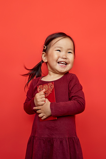 Vertical portrait of happy Asian girl holding hongbao envelope and smiling with candid emotion standing against red background