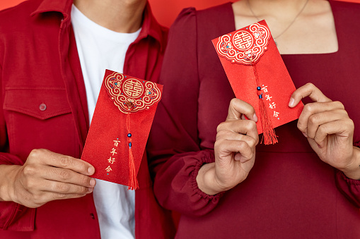 Close up of young Chinese couple holding red hongbao envelopes, copy space Translation Have overflowing abundance hundred years