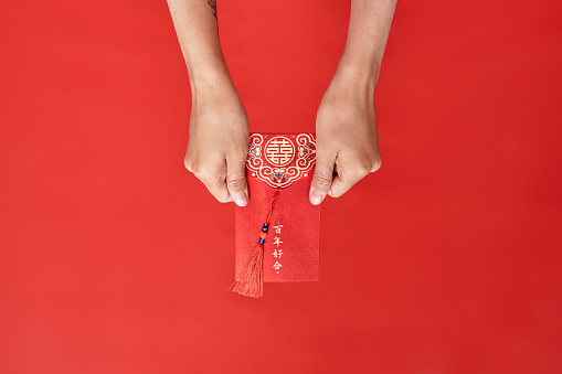 Close up of female hands holding hongbao envelope against red background as Chinese new year tradition, copy space Translation Have overflowing abundance hundred years