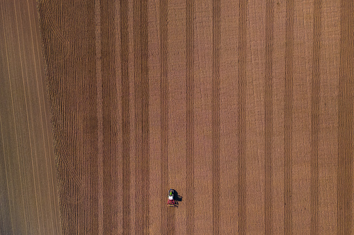 A small lonely tractor with plow on a large brown field with plowed earth seen directly from above