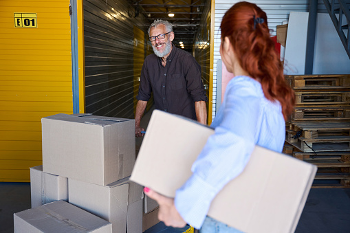 Bearded man and a red-haired lady are transporting boxes with things on a cargo cart, people are in a warehouse
