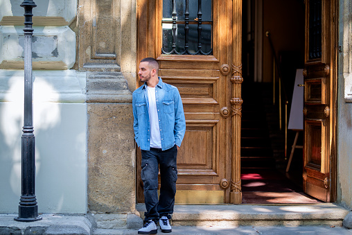 Full length portrait of a stylish young man standing in the doorway in a city street. Young male looking away and wearing casual clothes.