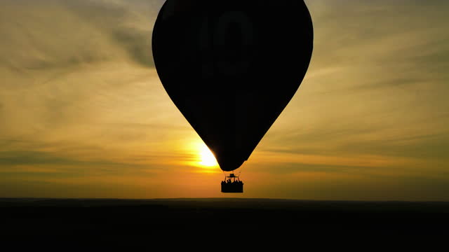 Aerial drone view of silhouette hot air balloon flying against sun at summer sunrise. Balloon hot air helium sunset sunrise. Establishing shot of aerial flight over hot air balloon